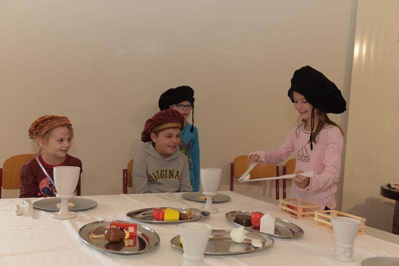 Szenisches Spiel: Zwei Kinder sitzen am gedeckten Tisch, während ein drittes Kind sie bedient.